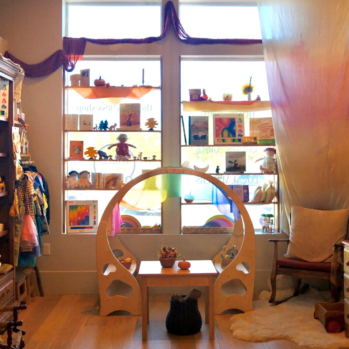 A cozy, whimsical children's room or play corner featuring a light wood arched play structure with a colorful rainbow hanging in the center, a small matching wooden table, and a large window in the background filled with shelves of colorful toys and books.
